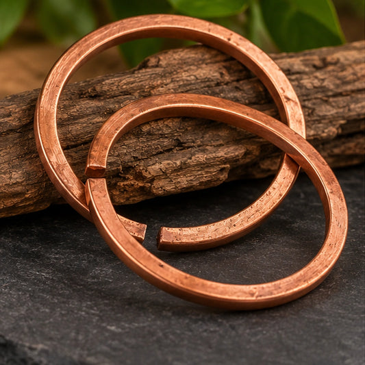 Deux bracelets ouverts en cuivre pur entrelacés devant une écorce de bois, sur une ardoise sombre entourée de verdure.
